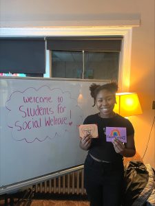 Student holding two pink cards, in front of a white board that says "Welcome Students for Social Welfare"