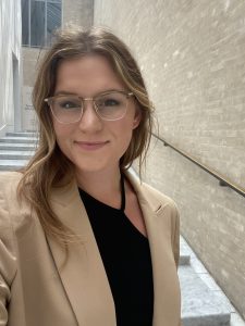 Haley in front of a brick stairwell, wearing a tan blazer, black dress, and glasses.