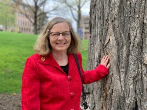 Margie Zutter in red coast standing outside with her hand against a large tree.